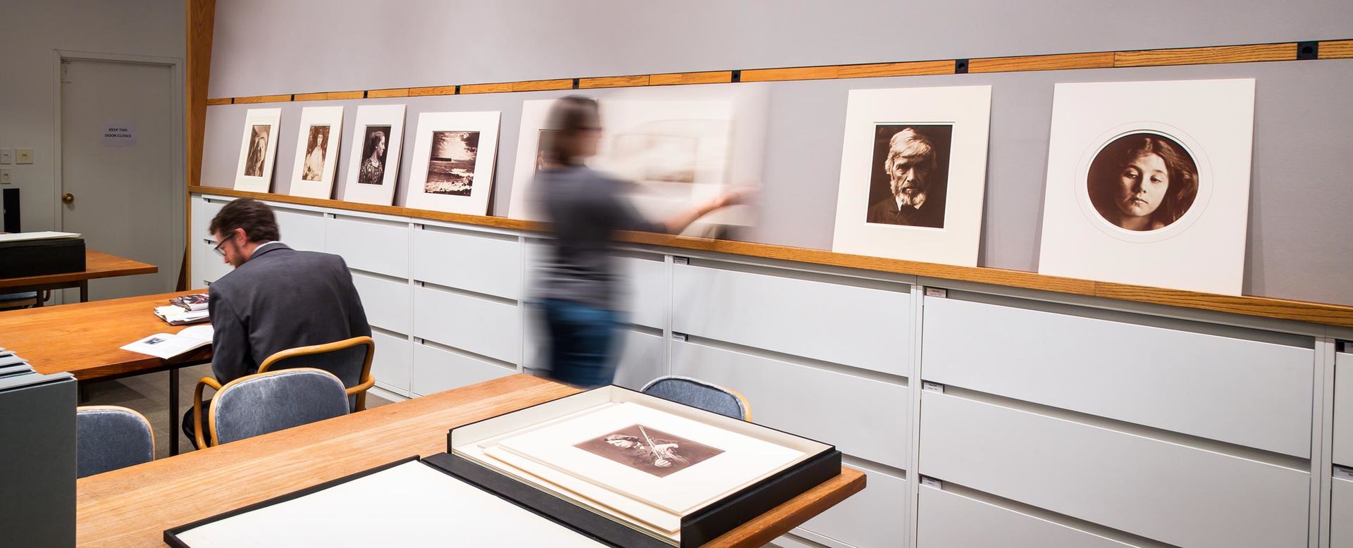 An office with blond wood tables and chairs, and against the back wall, low filing cabinets with a picture rail above for displaying photographic works of art